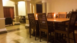Un comedor con una mesa redonda y seis sillas de madera, una vela sobre la mesa, y vistas a un pasillo con una puerta y una ventana con cortinas.