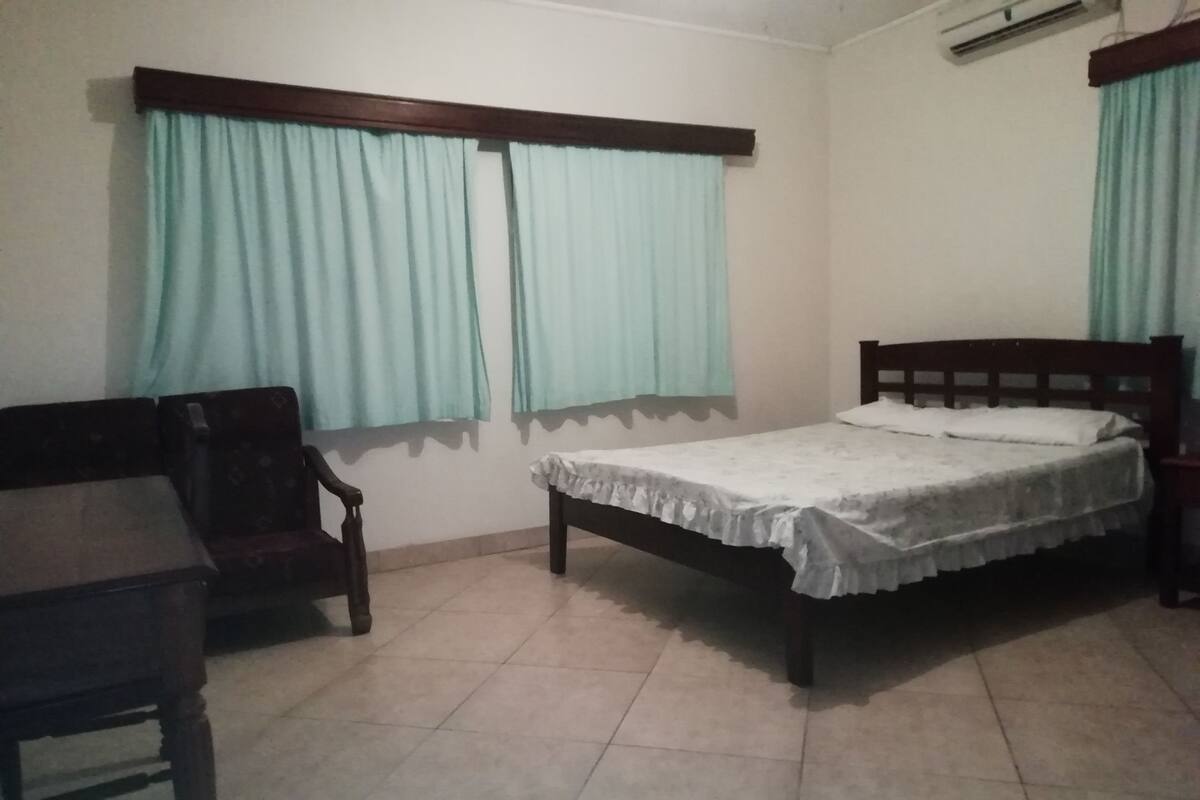 A bedroom with a single bed, a wooden chair, a small table, and light blue curtains.