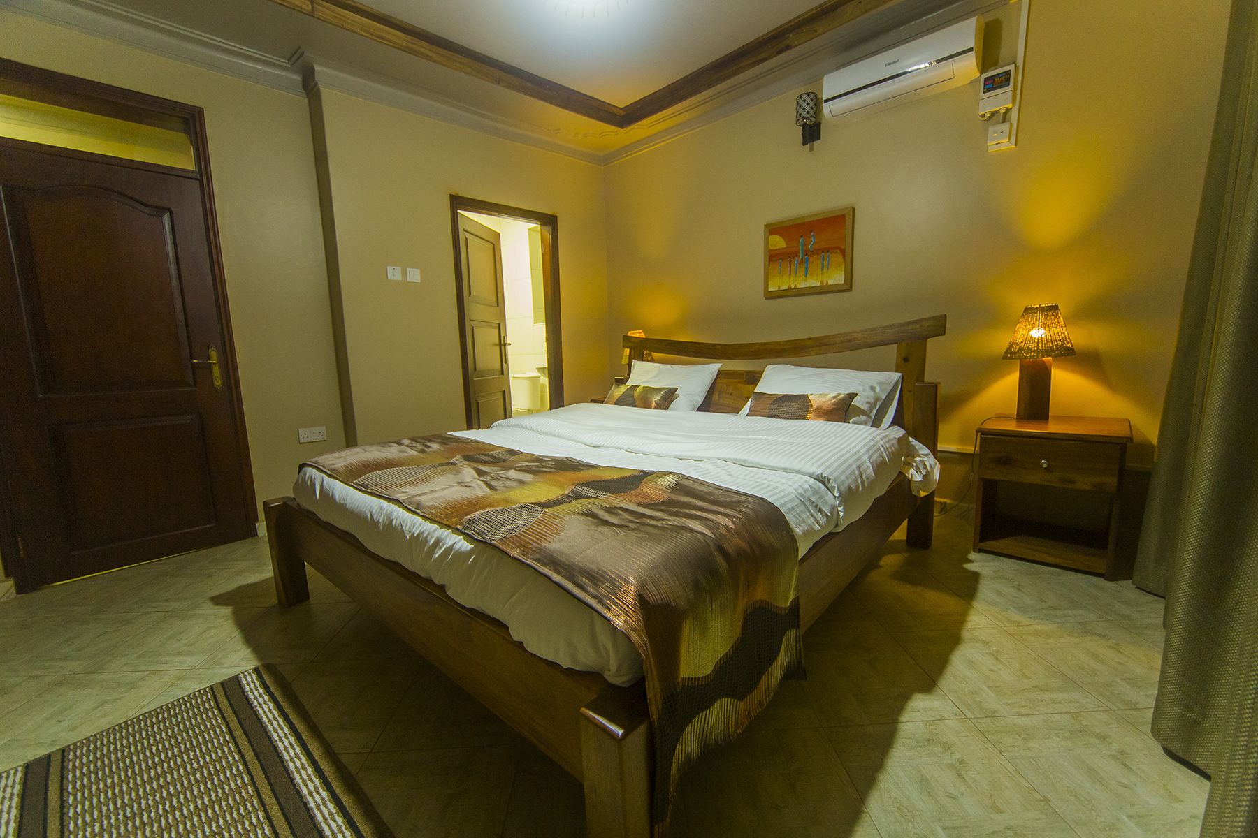 A hotel room with a large bed, a bedside table with a lamp, a painting on the wall, and a door.
