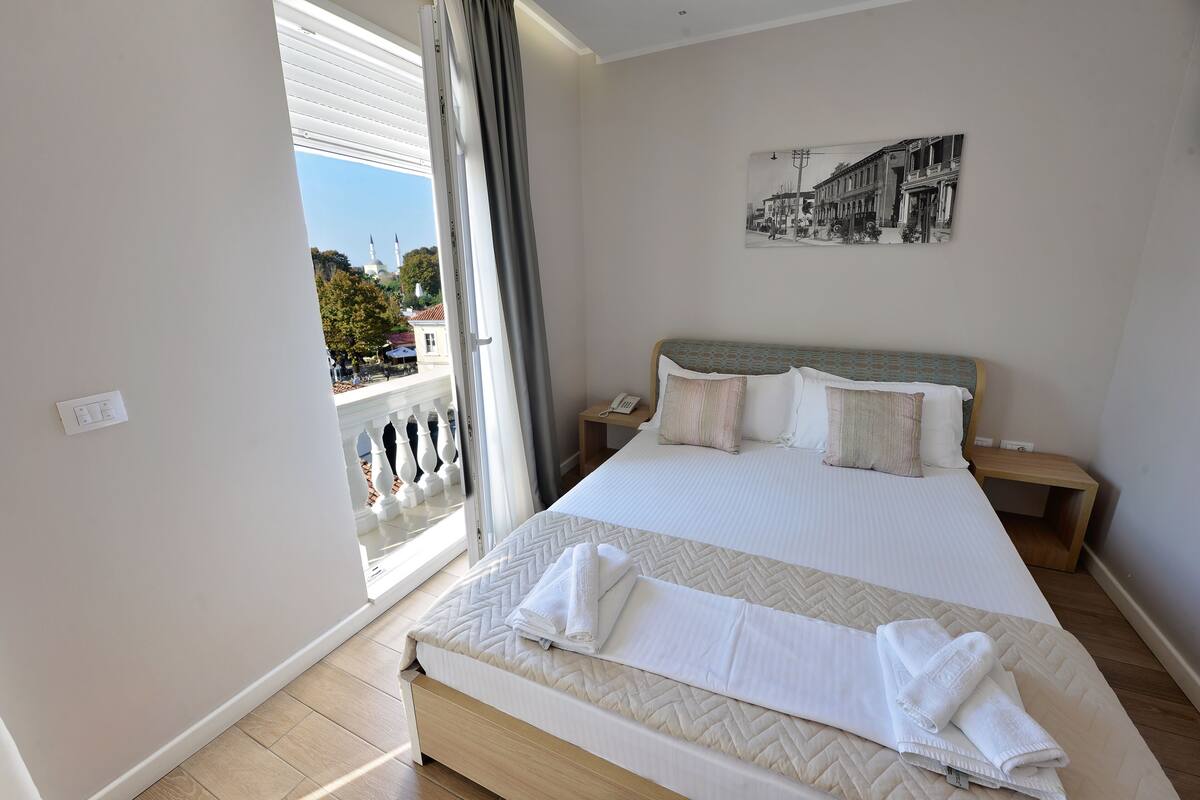A bedroom with a large bed, a view of a balcony, and a cityscape painting on the wall.