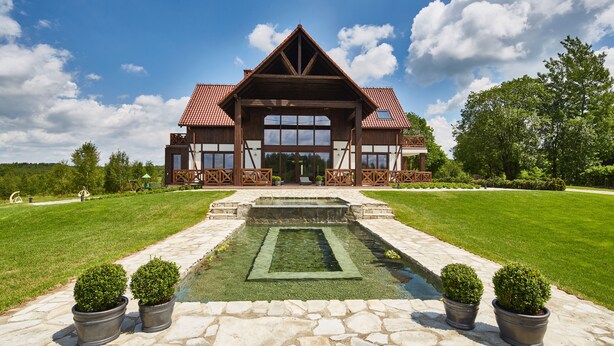 A large, two-story house with a prominent front porch, a stone pathway leading to a rectangular water feature, and a well-maintained lawn. - Siedlisko 518 Wzgorza Dylewskie (Ostróda)