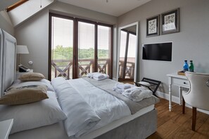 Une chambre avec un grand lit, un bureau, une chaise et une télévision. Il y a des fenêtres avec des stores et un balcon à l’extérieur.