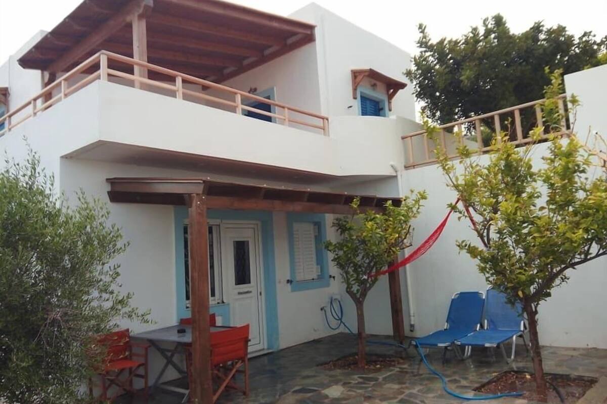 Un bâtiment à deux étages doté d’un balcon, d’un petit jardin avec un arbre et des chaises bleues, ainsi qu’une terrasse en pierre.