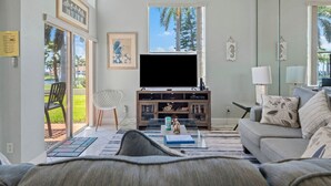 A modern living room with a large TV, comfortable seating, and a view of palm trees.