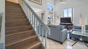 A spacious living room with a staircase, a flat-screen TV, and a ceiling fan.