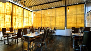 Un salón de conferencias con mesas y sillas, ventanas amplias con persianas y lámparas colgantes.