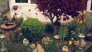 Un jardín con un bebedero rústico para pájaros, un árbol de hojas rojas y diversas plantas y flores.