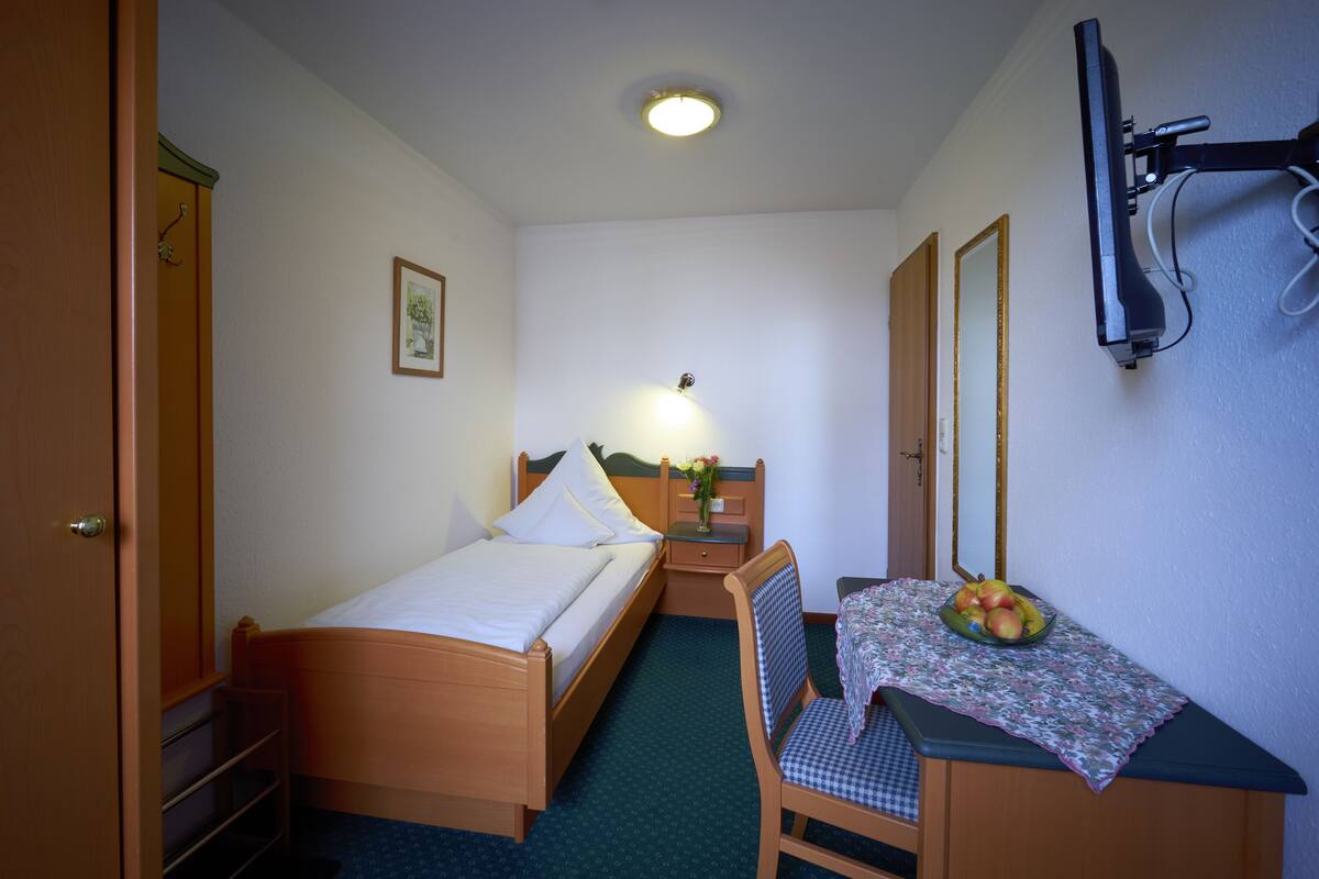 Ein kleines, ordentlich eingerichtetes Schlafzimmer mit einem Einzelbett, einem Sessel, einem kleinen Tisch mit einer Obstschale und einem Spiegel an der Wand.
