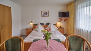 Habitación de hotel con cama, dos sillas, una mesa con un florero y televisión en la pared, además de una ventana con cortinas.