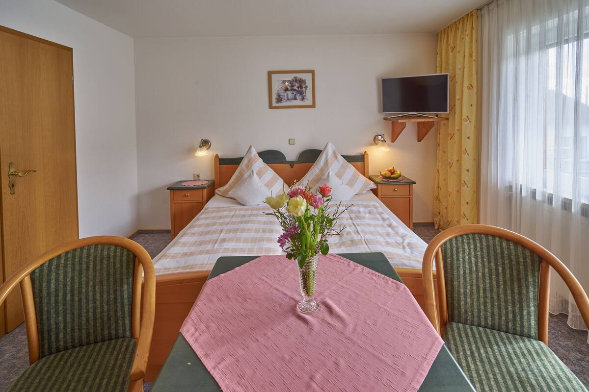 Ein Hotelzimmer mit Bett, zwei Stühlen, einem Tisch mit Blumenvase, einem an der Wand befestigten Fernseher und einem Fenster mit Vorhängen.