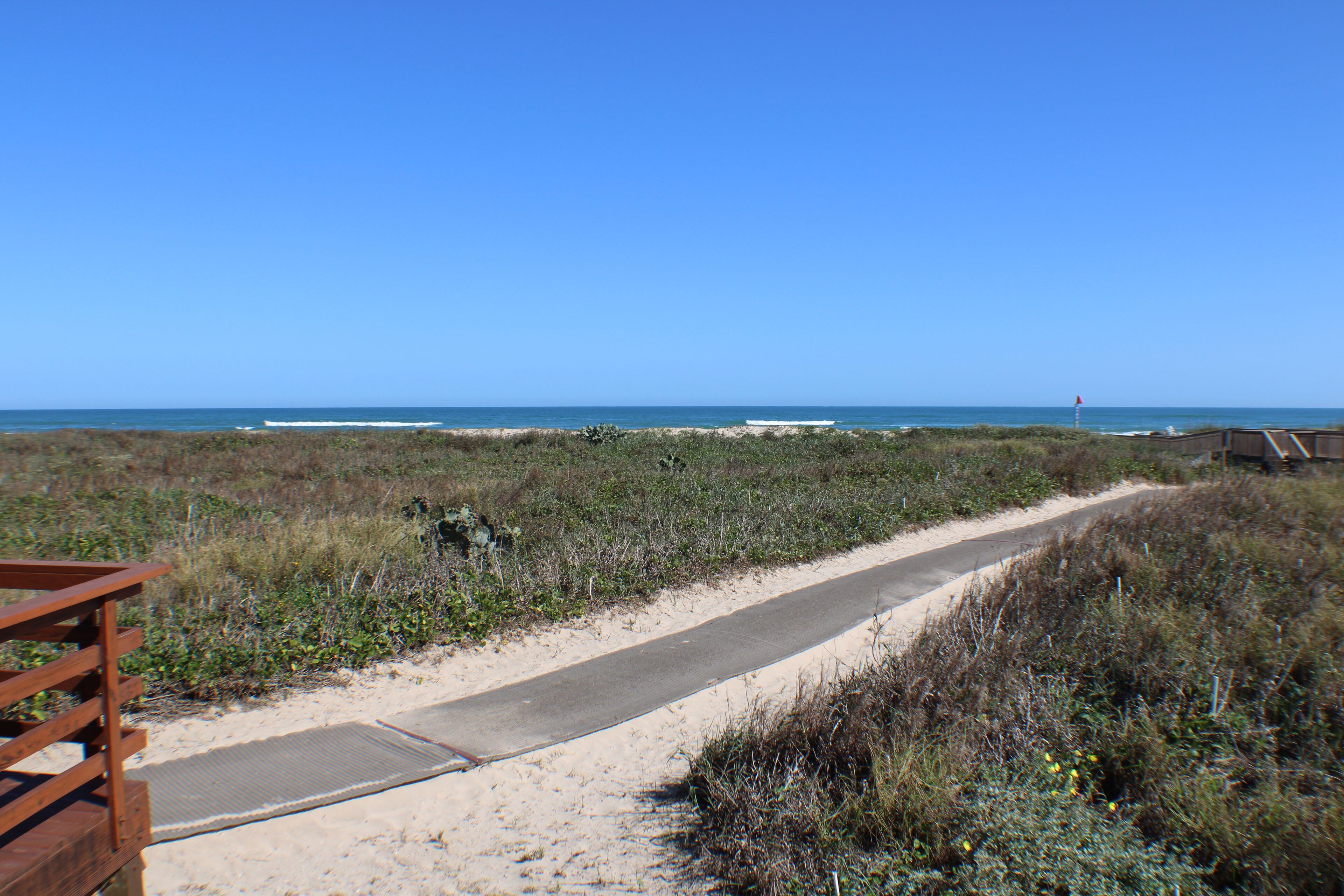 Un sentier bordant la plage, avec une rambarde en bois sur la gauche, qui mène à l’océan.