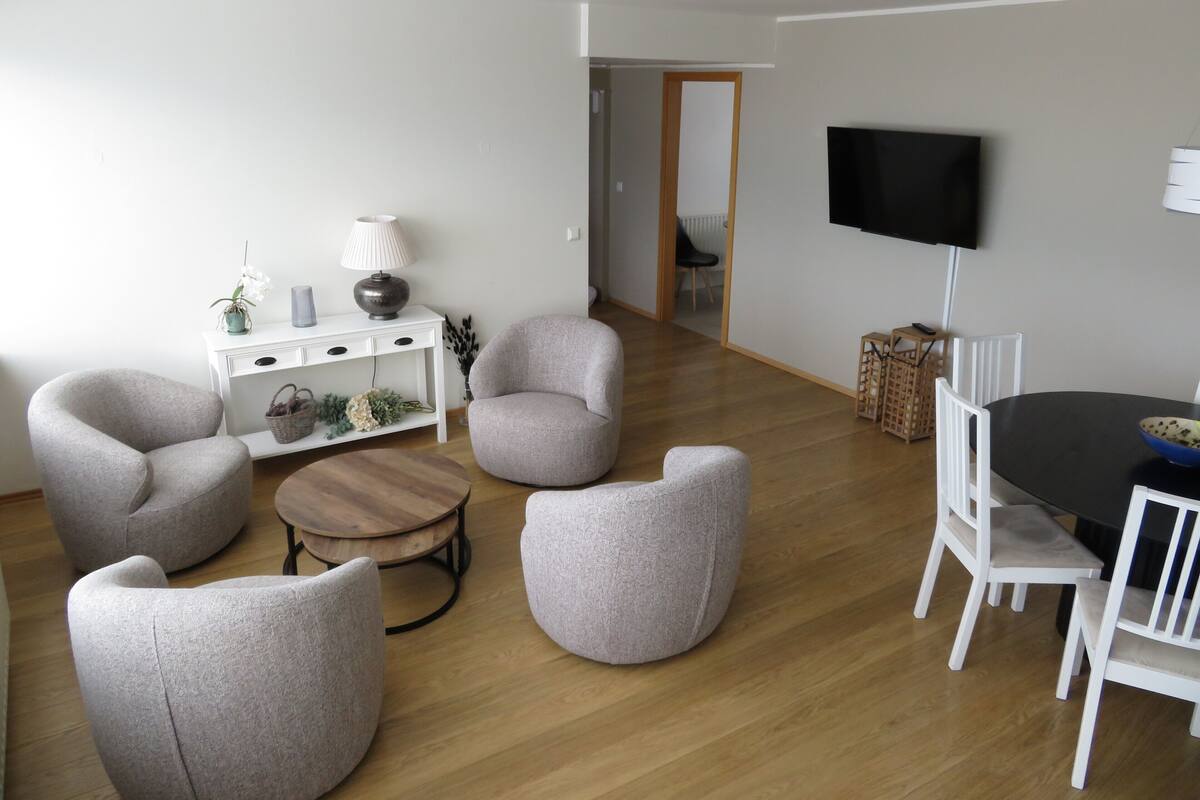 Una sala de estar con una mesa redonda de madera, sillones grises, una mesita de noche blanca con una lámpara, y un televisor montado en la pared.