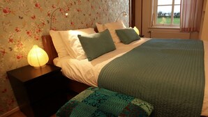 A bedroom with floral wallpaper, a bed with green and white bedding, a bedside lamp, and a window with curtains.