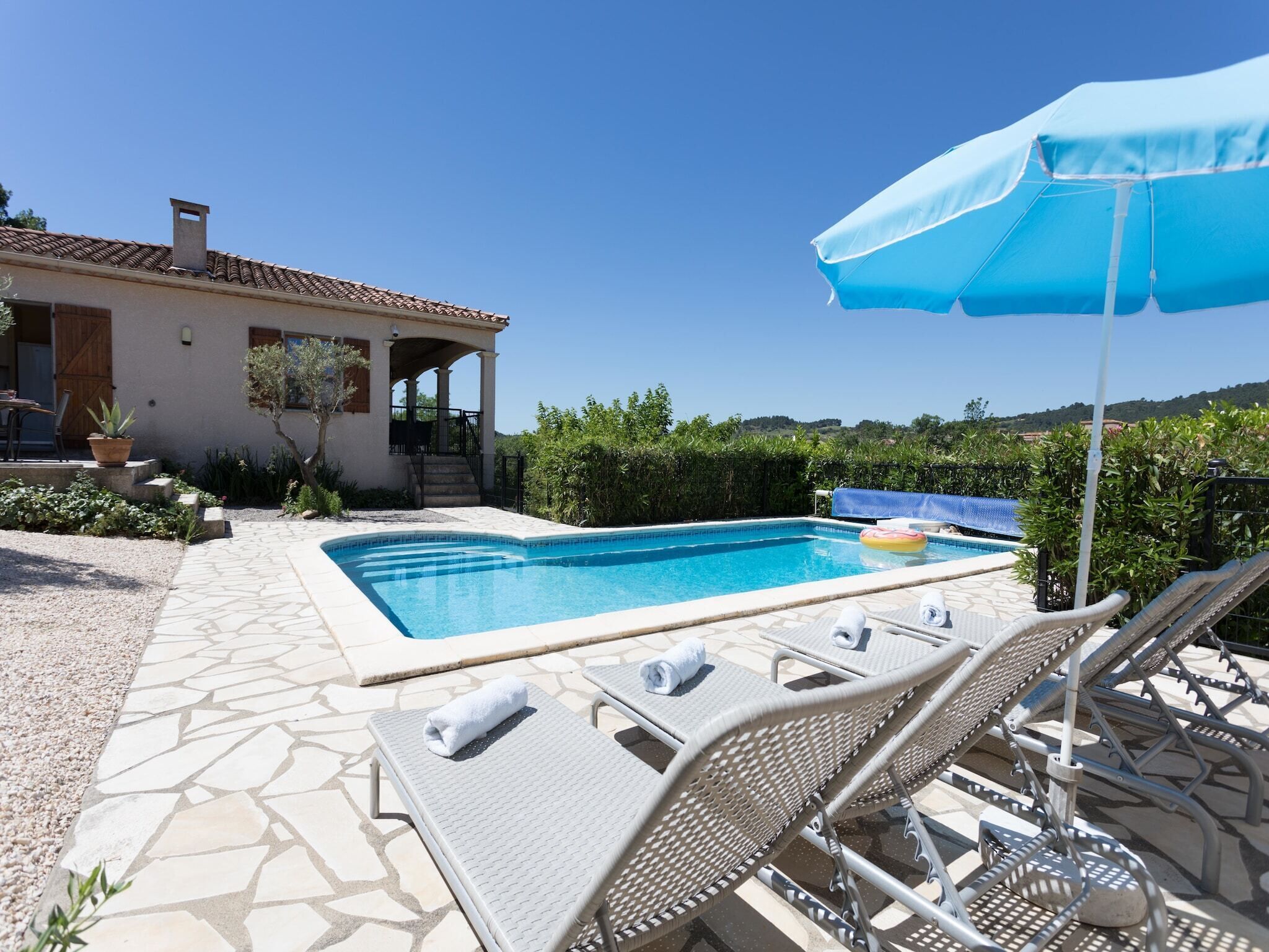A pool area with lounge chairs and an umbrella, surrounded by a stone patio and a building with a tiled roof.