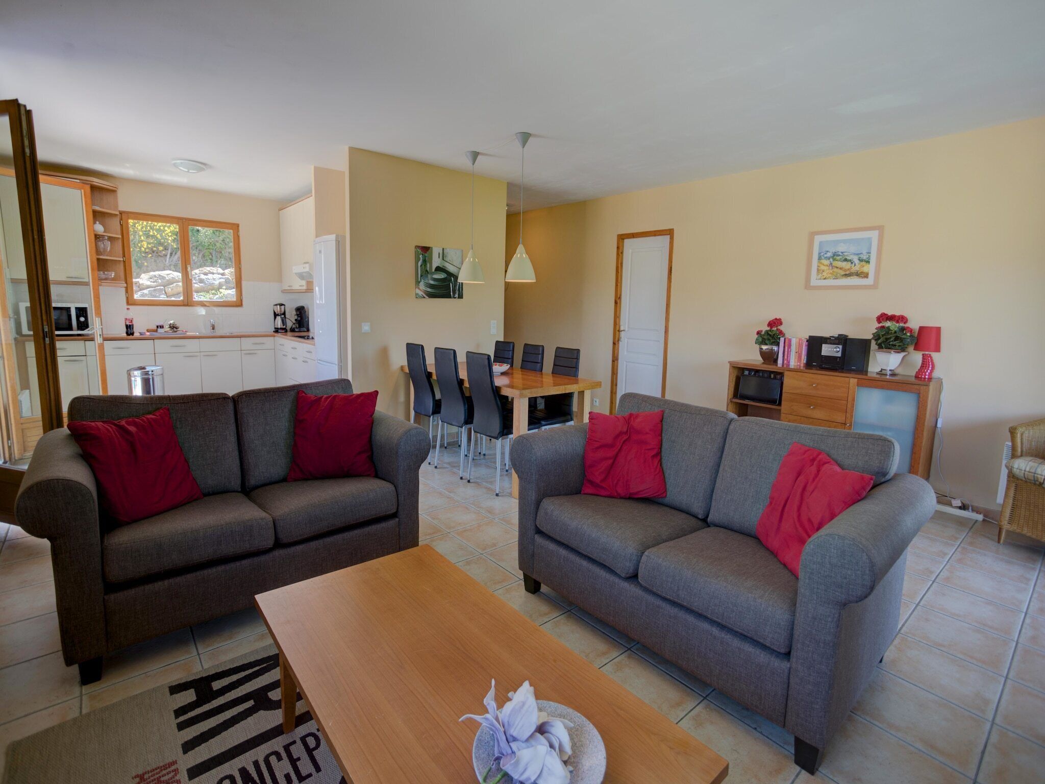 Un espacio habitable compacto con dos sofás grises, una mesa de centro de madera, un comedor con mesa y sillas de madera, una cocina con armarios blancos y un televisor pequeño sobre un mueble de madera.