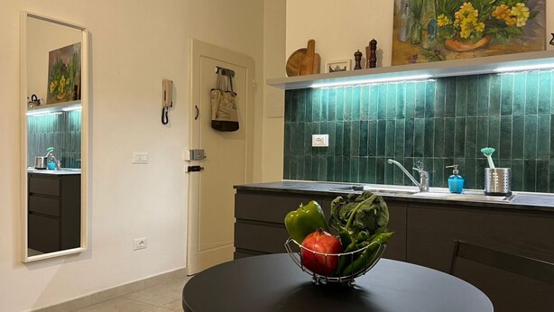 A modern kitchen with a round table, a fruit bowl, a green tiled backsplash, and a painting above the sink.
