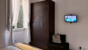 A bedroom with a wardrobe, a bed, a TV mounted on the wall, and a window with curtains.