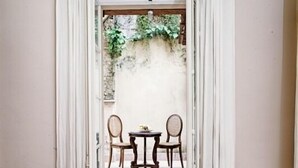 Quarto de hotel com tapete vermelho, cortinas brancas e vista para uma área externa com dois cadeiras e uma mesa.