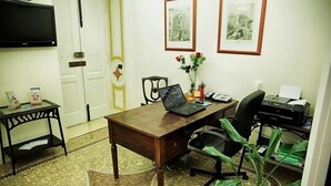 Um quarto com uma escrivaninha de madeira, um laptop, uma impressora, uma cadeira, uma TV fixada na parede e quadros enquadrados na parede.