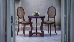 Uma porta aberta dá para um pátio com uma mesa e duas cadeiras de vime, uma pequena tigela de frutas na mesa, e uma parede de pedra com vegetação.
