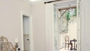 Quarto de hotel com colcha vermelha, cama com moldura dourada, pequena mesa de cabeceira com abajur e vista para um pátio exterior através de uma porta aberta.