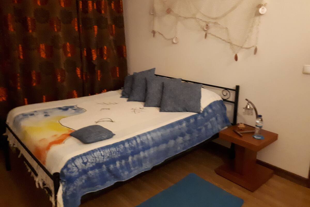 A bedroom with a bed, pillows, a nightstand, a lamp, and a curtain.