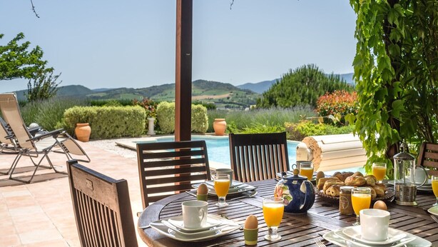 A dining table set for breakfast with a view of a pool and hills in the background.