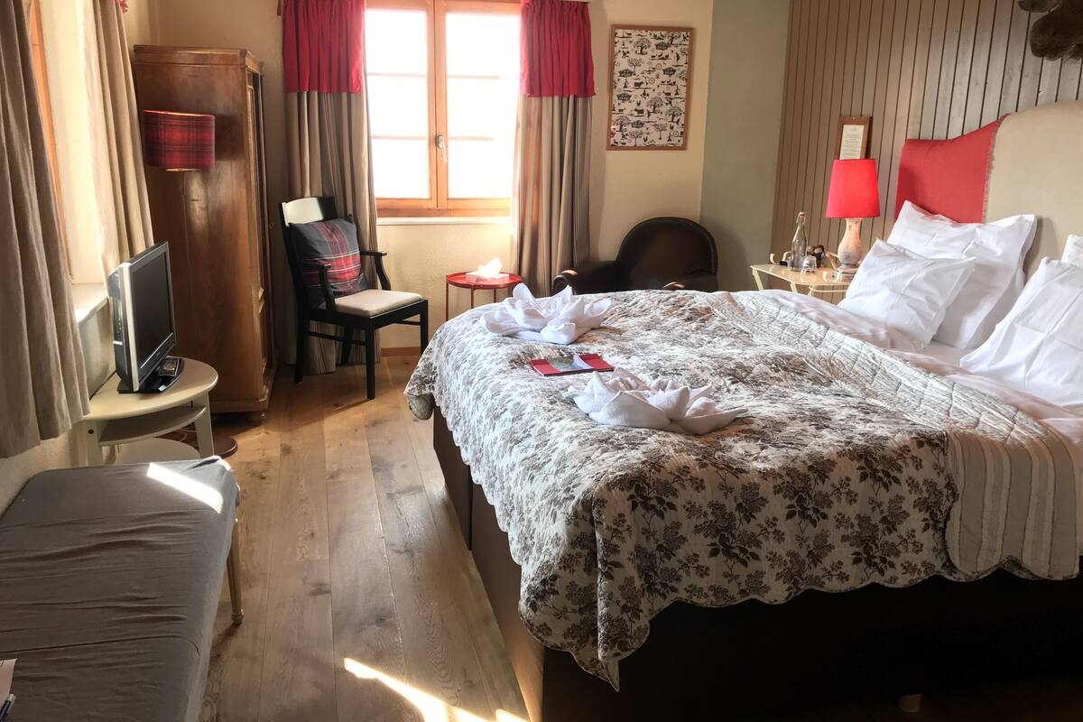 Una habitación de hotel con cama, escritorio con computadora, silla, mesita con lámpara y ventana con cortinas.