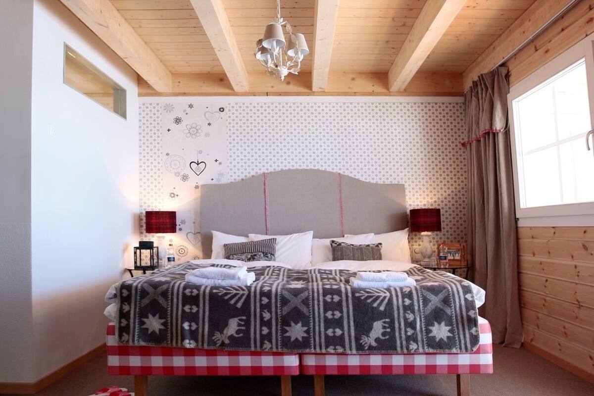 Un dormitorio con una cama grande, vigas de madera, un candelabro, un papel tapiz estampado y una ventana con cortinas.