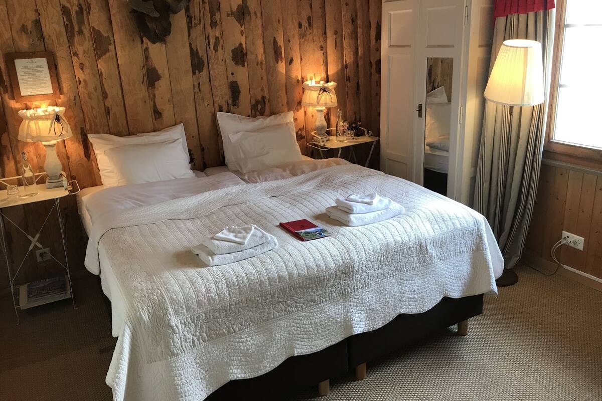 Una cama bien tendida con sábanas blancas, dos mesitas de noche con lámparas, una pared de madera y una ventana con cortinas.