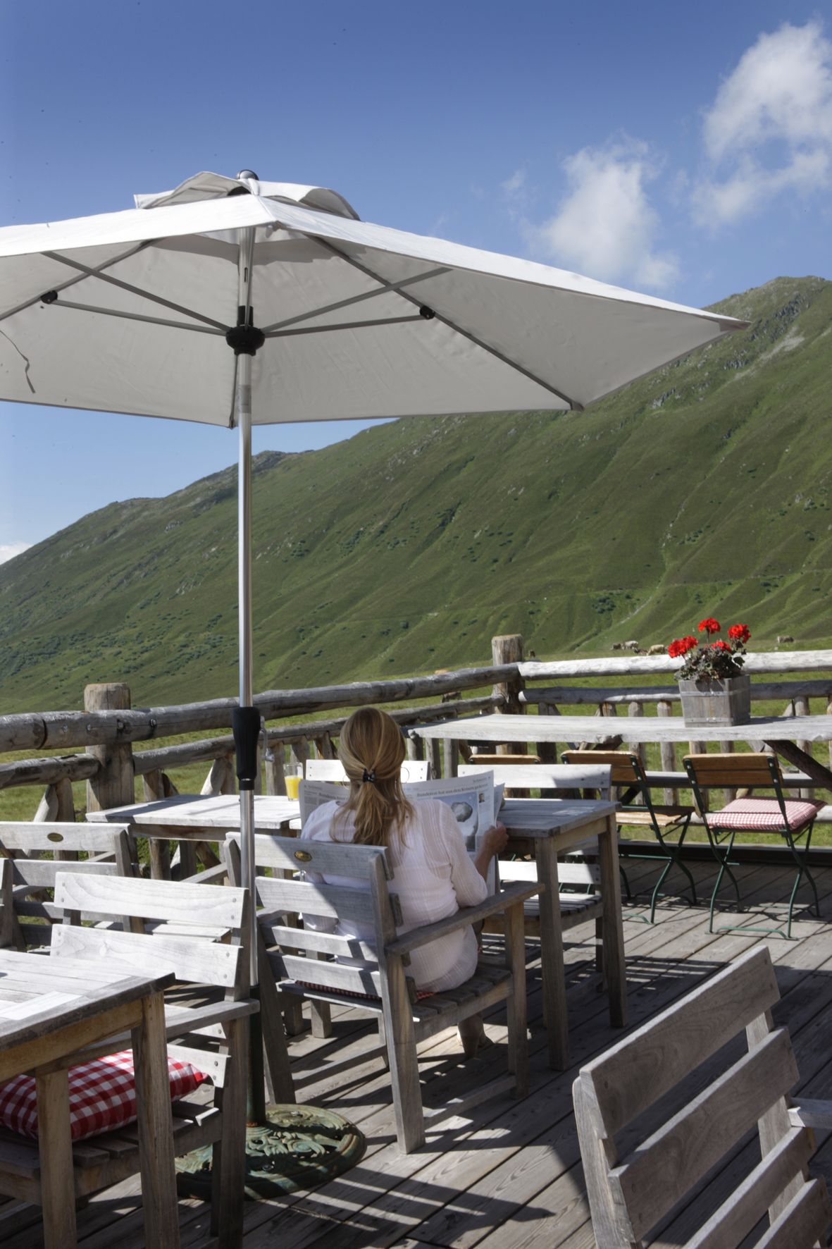 A woman reading a newspaper at a wooden table with a white umbrella, overlooking a mountainous landscape.