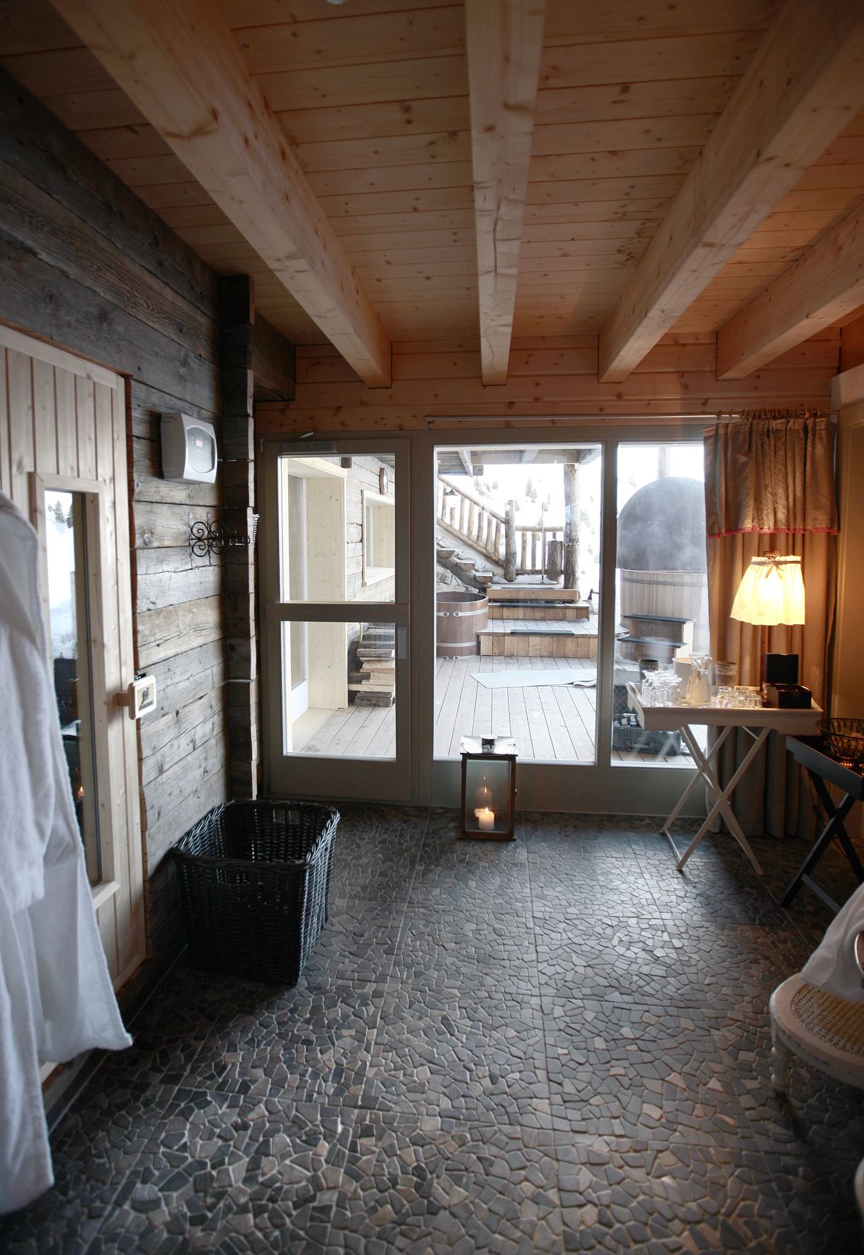 A cozy room with a stone-tiled floor, wooden walls, a large window with a view of a wooden deck, and a table with a lamp.