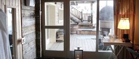 A cozy room with a stone-tiled floor, wooden walls, a large window with a view of a wooden deck, and a table with a lamp.