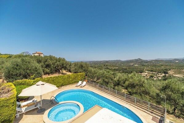 A swimming pool with two hot tubs, surrounded by a fence, and overlooking a hilly landscape with a clear sky.