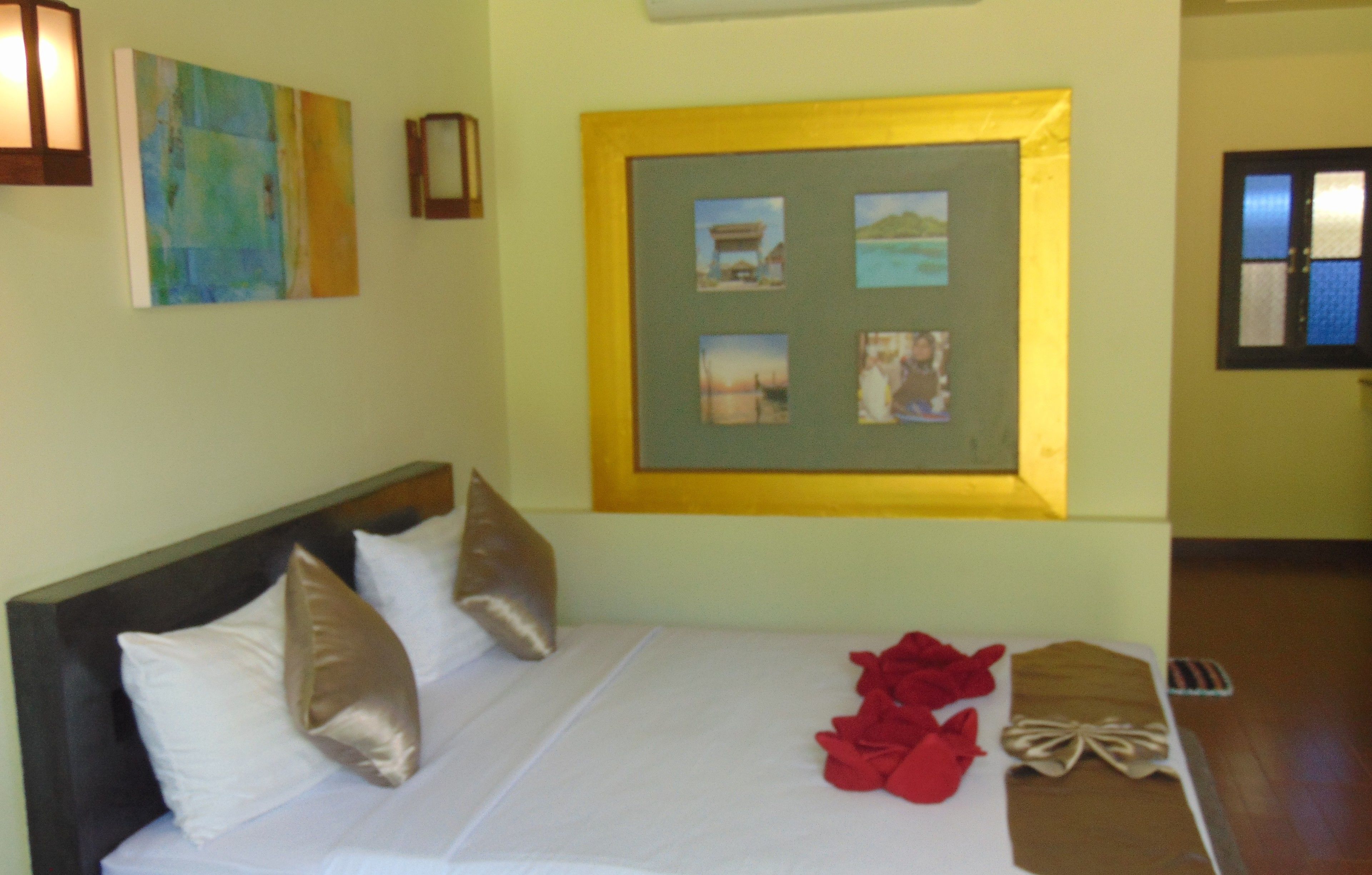 A neatly made bed with white and gold pillows, a framed picture on the wall, and a small table with red and brown items.