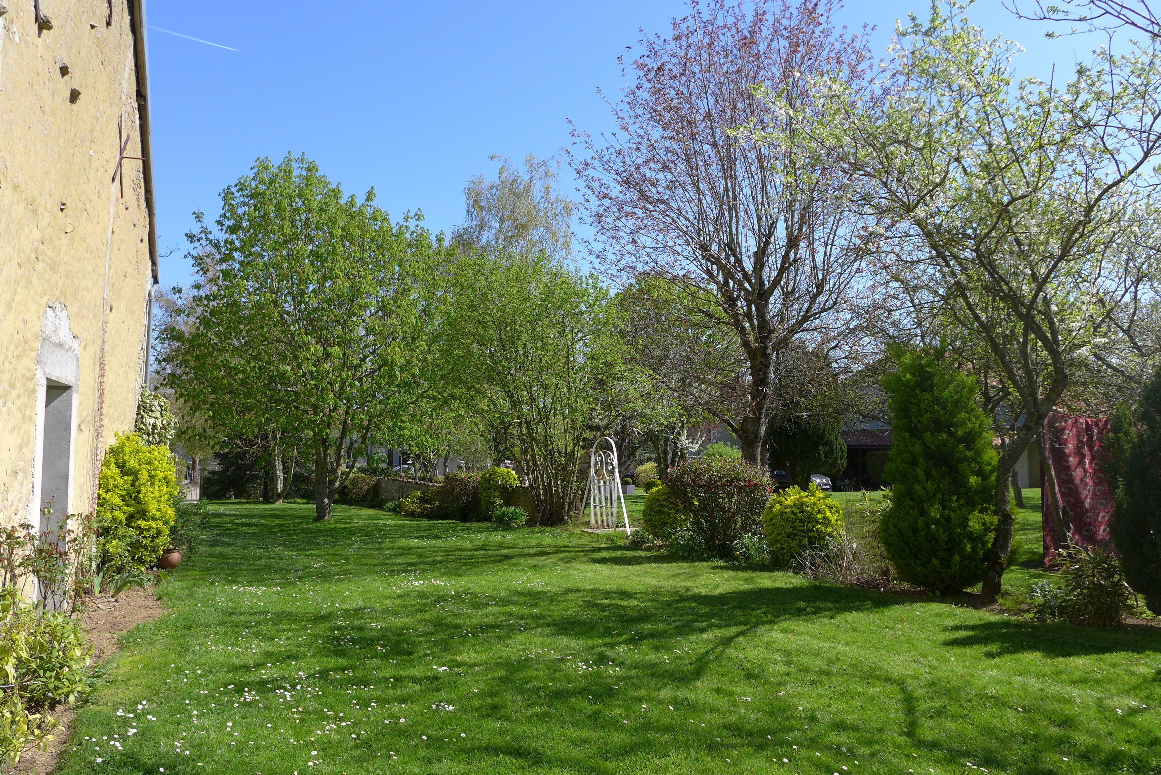 A well-maintained garden with a variety of trees and shrubs, a clear sky, and a building with a yellow facade.