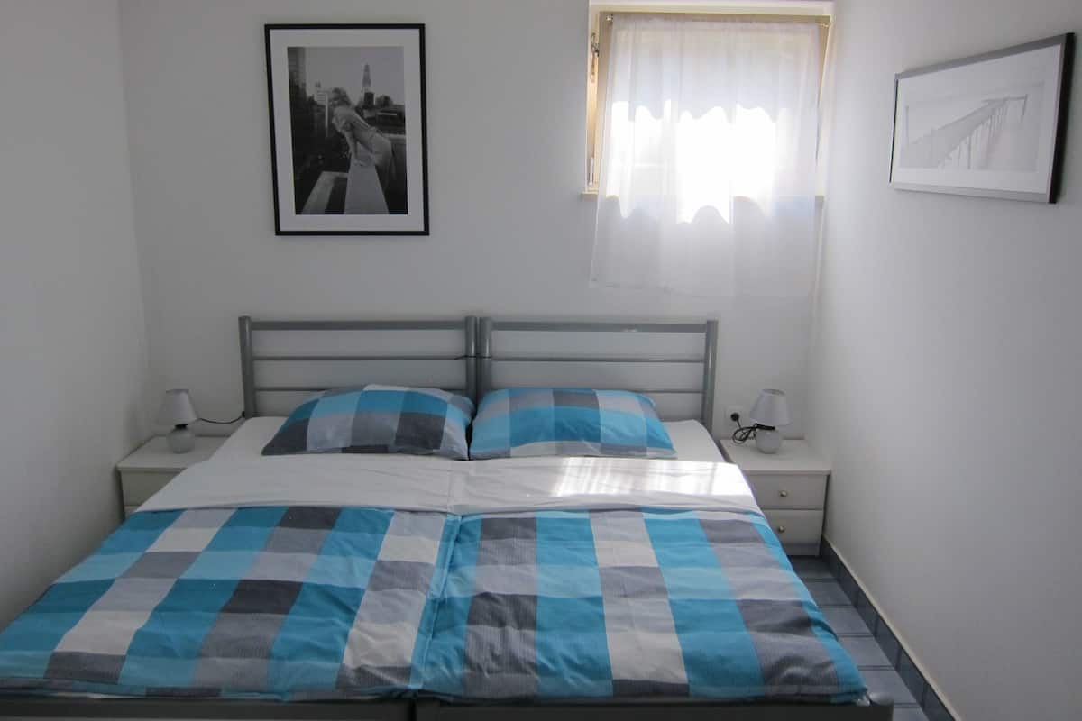 A bedroom with a bed, two bedside tables, a window with curtains, and two framed pictures on the wall.