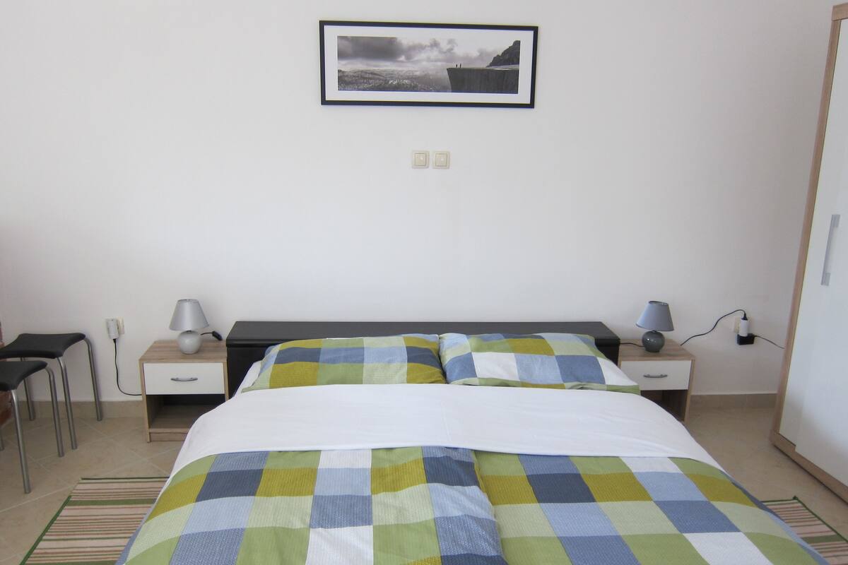 A bedroom with a bed, two bedside tables, a chair, a lamp, and a framed picture on the wall.