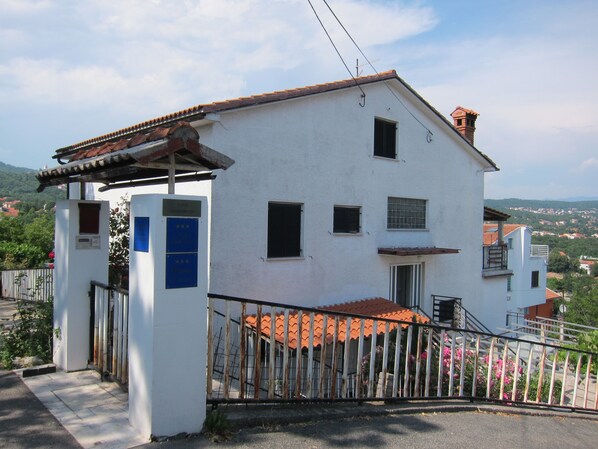 A white building with a red-tiled roof, a gated entrance, and a view of a hilly landscape in the background. - Nada Apartments (Opatija)