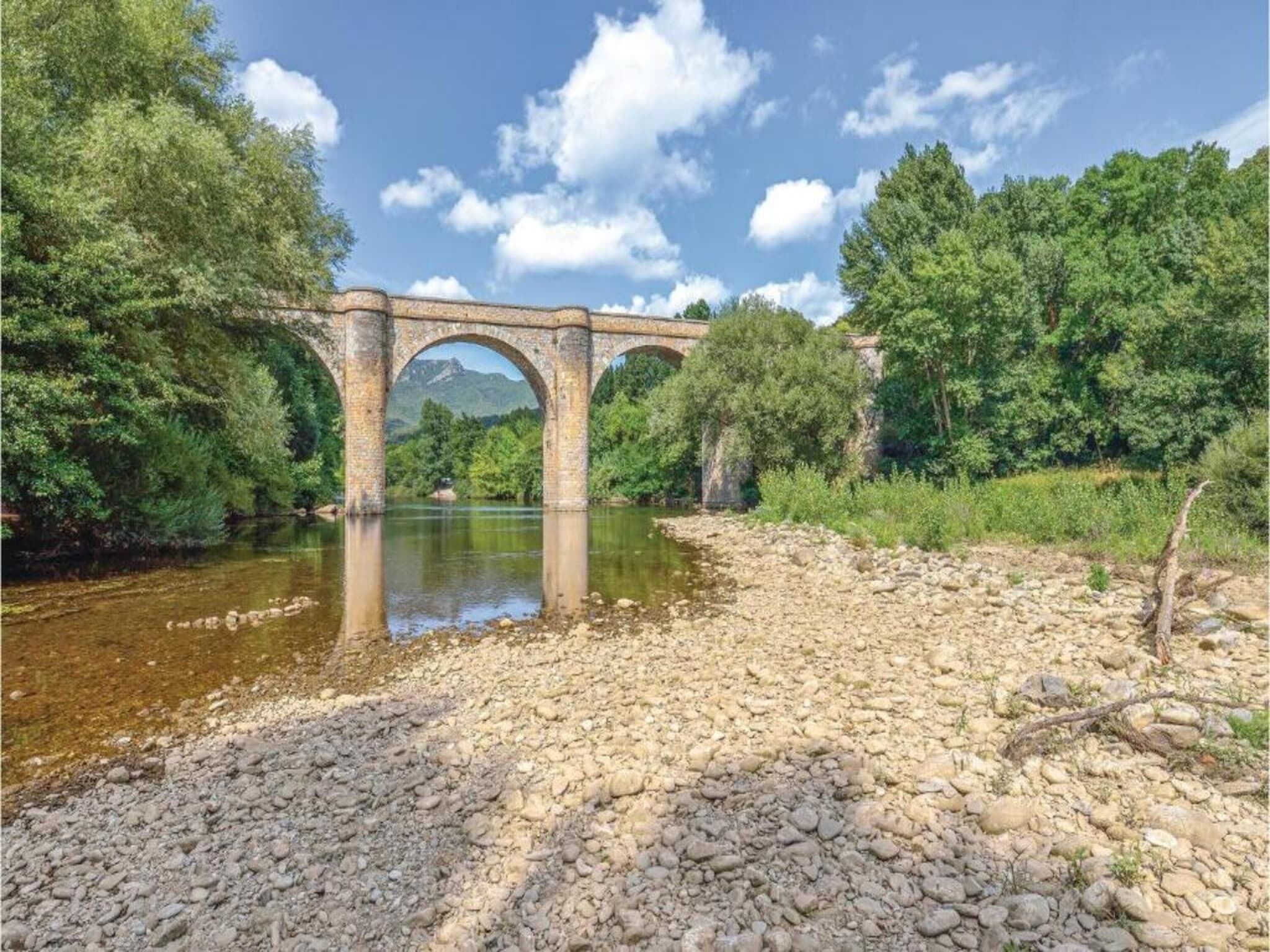 A stone bridge over a river with rocky banks and dense greenery on both sides.