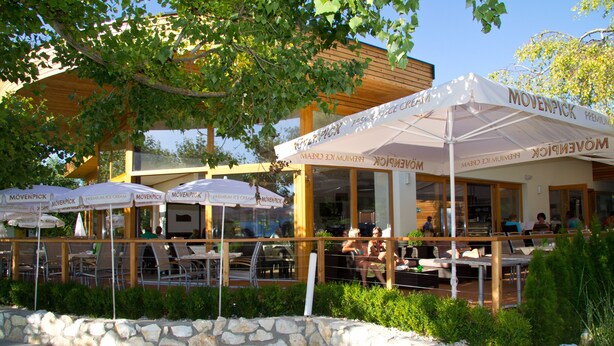 An outdoor seating area with tables and chairs under umbrellas, a stone wall, and a building with large windows. - Modern apartment with large balcony on Lake Klopein (St. Kanzian)