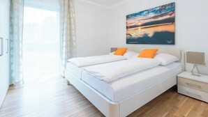 A double bed with white bedding and orange pillows, a bedside table with a lamp, and a large window with sheer curtains. - Modern apartment with large balcony on Lake Klopein (St. Kanzian)