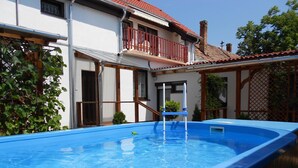 En swimmingpool med ett trädäck och ett hus med ett rött tegeltak i bakgrunden.
