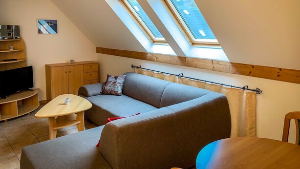 A loft-style room with a sloped ceiling, wooden beams, a grey sofa, a small table, and a TV. - Vacation apartment Ostseestern 19O-2 bedrooms up to max. 4 pers. + 2 baby - Holiday home (Trassenheide)