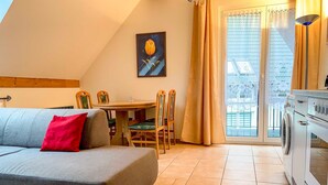 A cozy living area with a sofa, a dining table, and chairs. There is a painting on the wall, a window with curtains, and a washing machine. - Vacation apartment Ostseestern 19O-2 bedrooms up to max. 4 pers. + 2 baby - Holiday home (Trassenheide)