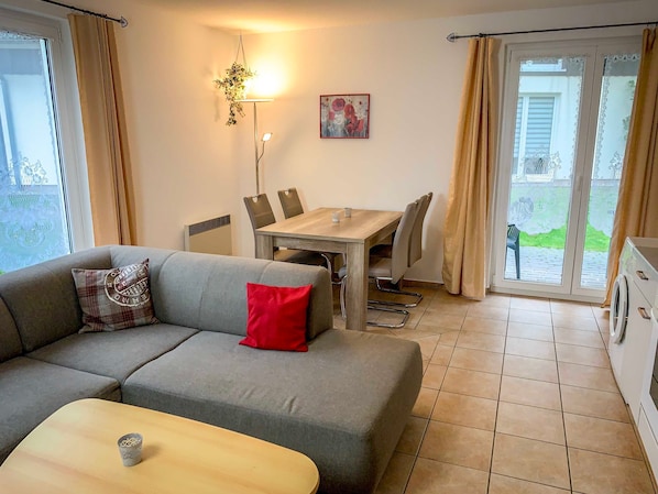 A living room with a grey sofa, a wooden coffee table, a dining table with chairs, and a kitchen area with a washing machine.