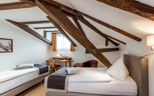A room with a sloped ceiling, wooden beams, a bed, a desk, a chair, and a lamp. - Romantik Hotel Stern (Chur)