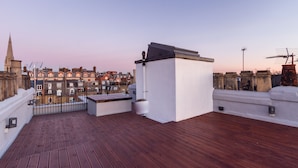 Eine Dachterrasse mit Holzboden, einer kleinen weißen Struktur und Blick auf eine Häuserreihe.