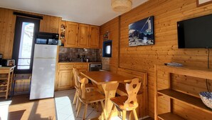 Una cocina con armarios de madera, una mesa de comedor y sillas. Hay un refrigerador, un lavaplatos y una estufa. Un televisor está montado en la pared.