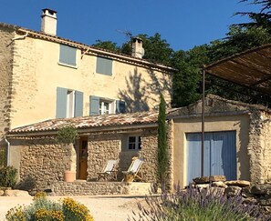 A stone building with a tiled roof, a chimney, and a small porch with two chairs and a table.